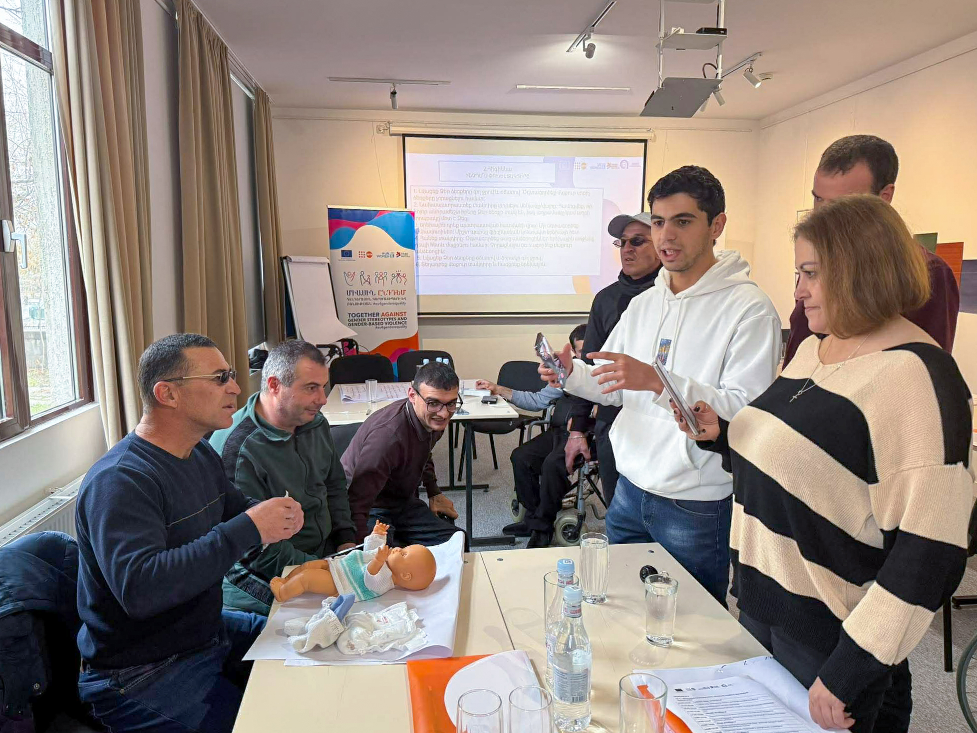 լուսնկարում 7 անձ է, հավաքված սեղանի շուրջ, մասնակիցներից մեկի դիմաց դրված է տիկնիկ, ցույց է տալիս, ինչպես կարելի է փոխել երեխայի տակդիրը