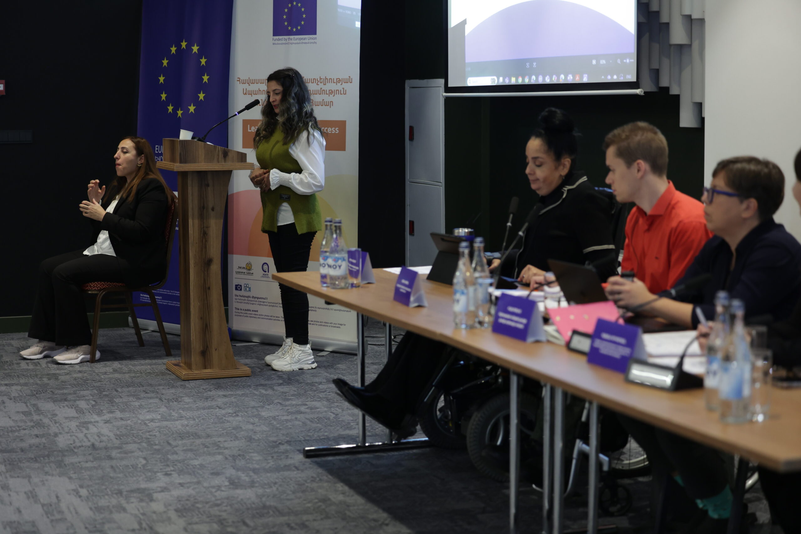 Լուսնկարում կոնֆերանսի հաղորդավարն է կանգնած ամբիոնի մոտ, կողքին՝ ժեստերի լեզվի թարգմանիչն է, սեղանի դիմաց նստած են եվրոպական անկախ կյանքի ցանցի 3 ներկայացուցիչները